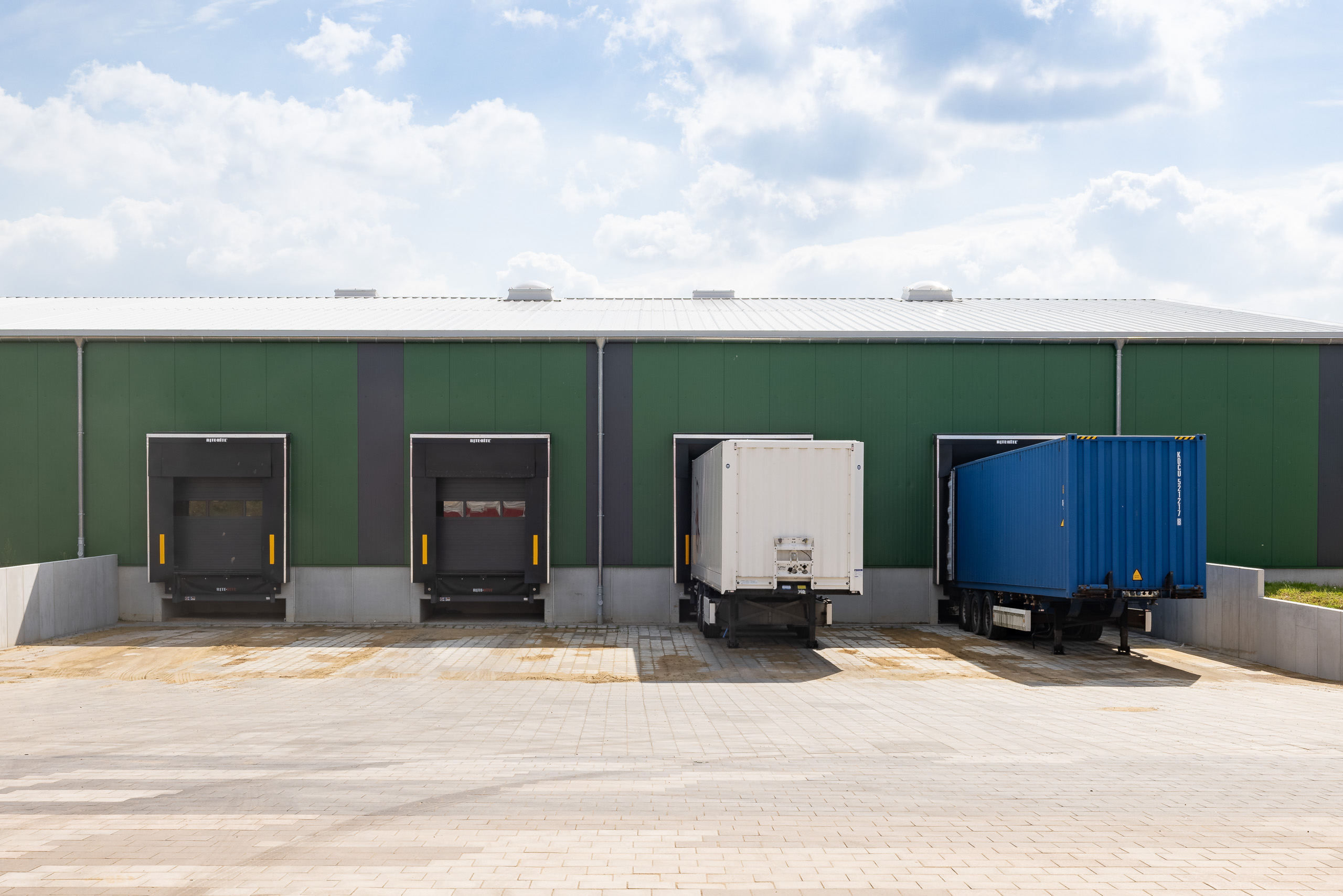 Ladezone einer grünen Halle mit vier Lademöglichkeiten. Zwei Ladezonen sind mit einem Containeranhänger besetzt, rechts ein blauer Anhänger und Mitte rechts ein weißer Anhänger.